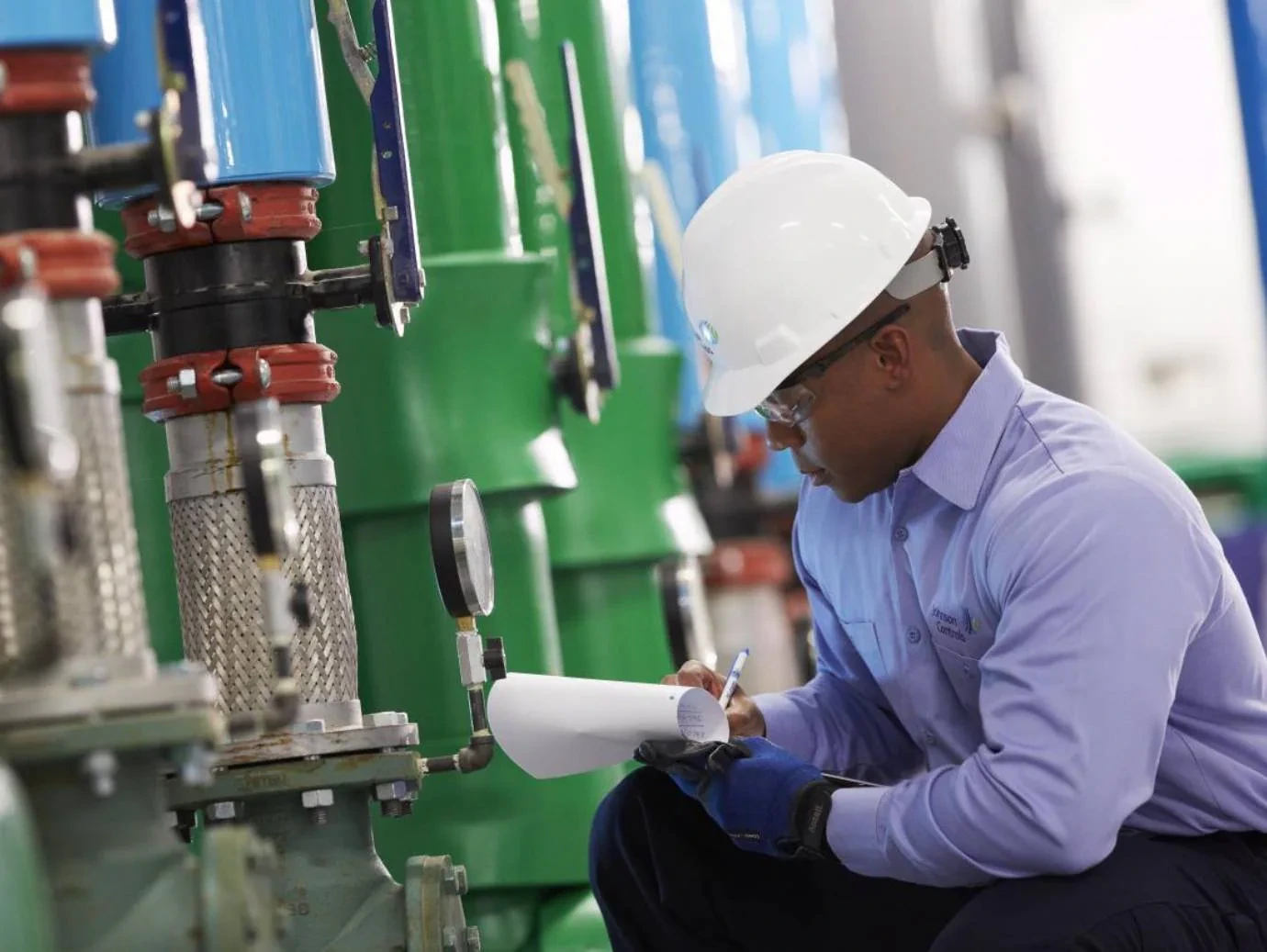 Mechanic in a white hard hat recording equipment readings beside pipes and a pressure gauge