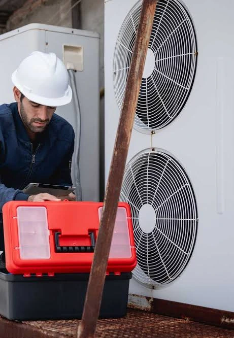 HVAC technician in a white hard hat inspecting an outdoor cooling system beside a red toolbox